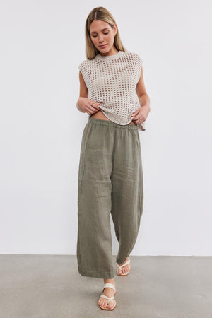 A person stands against a white background wearing a sleeveless beige top and Velvet by Graham & Spencer's LOLA LINEN PANT with an elastic waist, paired with light sandals. They are looking down and slightly adjusting their top.