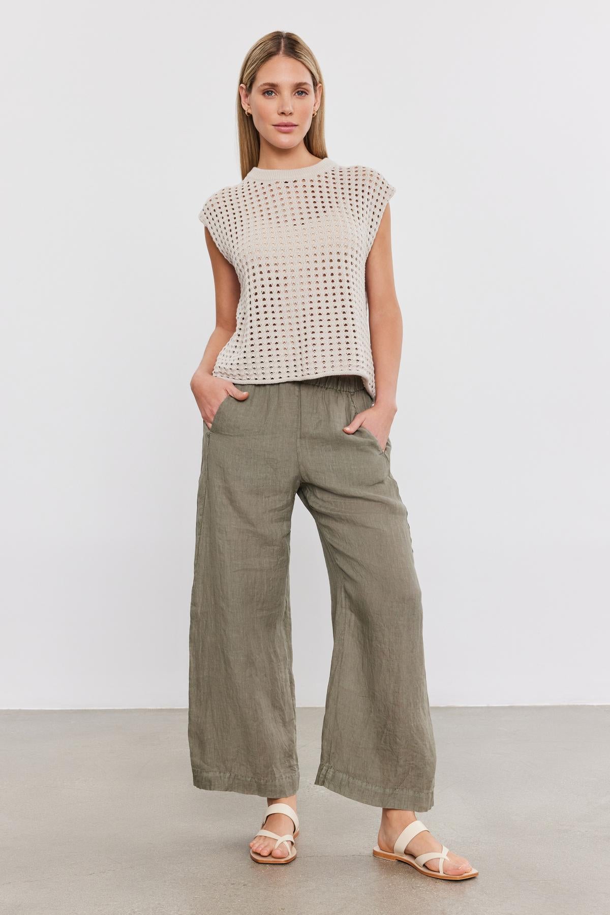 A person stands against a plain background, wearing a light beige knitted sleeveless top, LOLA LINEN PANT by Velvet by Graham & Spencer in olive green, and white sandals. Hands are in pockets, and the person is looking straight ahead.-36910043070657