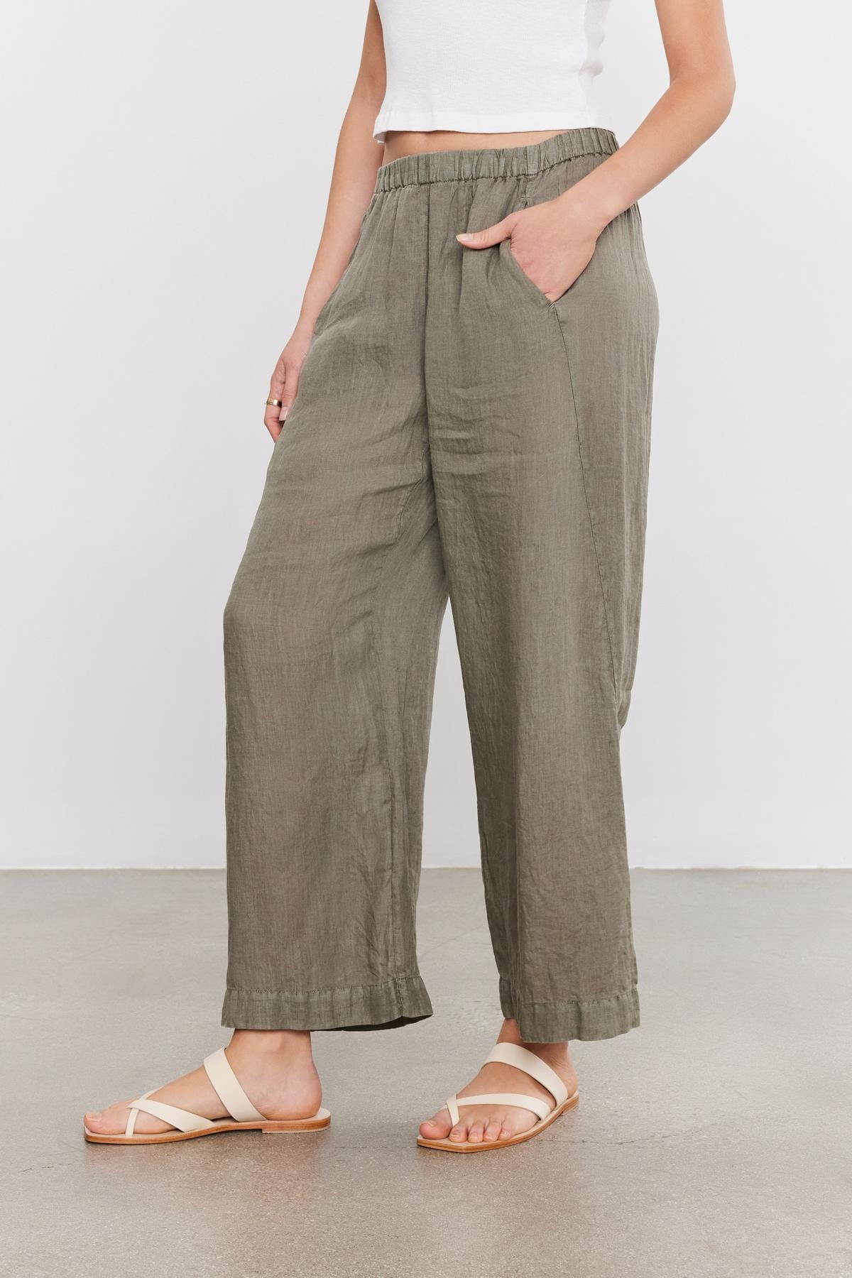   A person wearing olive green LOLA LINEN PANT by Velvet by Graham & Spencer with hands in pockets, a white crop top, and beige sandals. 