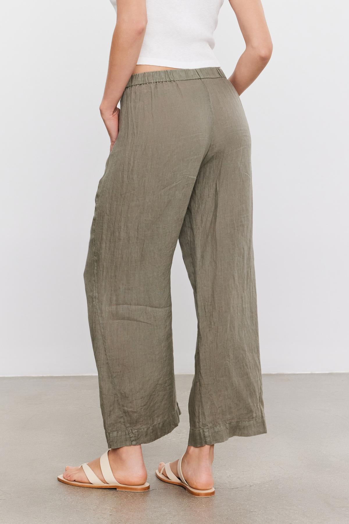   Rear view of a person wearing loose-fitting, lightweight LOLA LINEN PANT by Velvet by Graham & Spencer in olive green, a white top, and beige sandals, standing against a plain white background. 