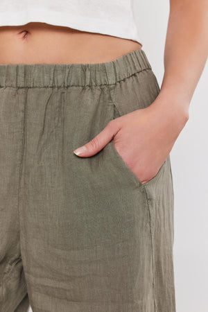 A person wearing green, relaxed leg LOLA LINEN PANT by Velvet by Graham & Spencer and a white top has their right hand in their pants pocket.