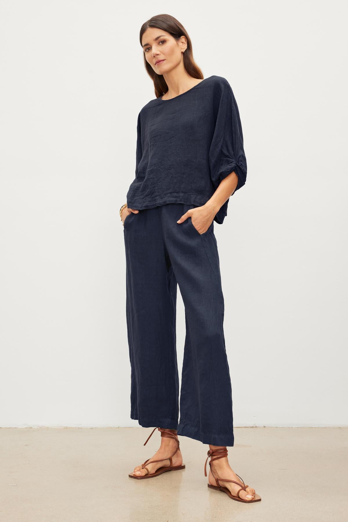 A woman in a loose-fitting Velvet by Graham & Spencer CASS LINEN CREW NECK TOP with a cocoon silhouette and wide-leg pants stands with hands in pockets. She is wearing brown sandals and has long hair.-37240045633729