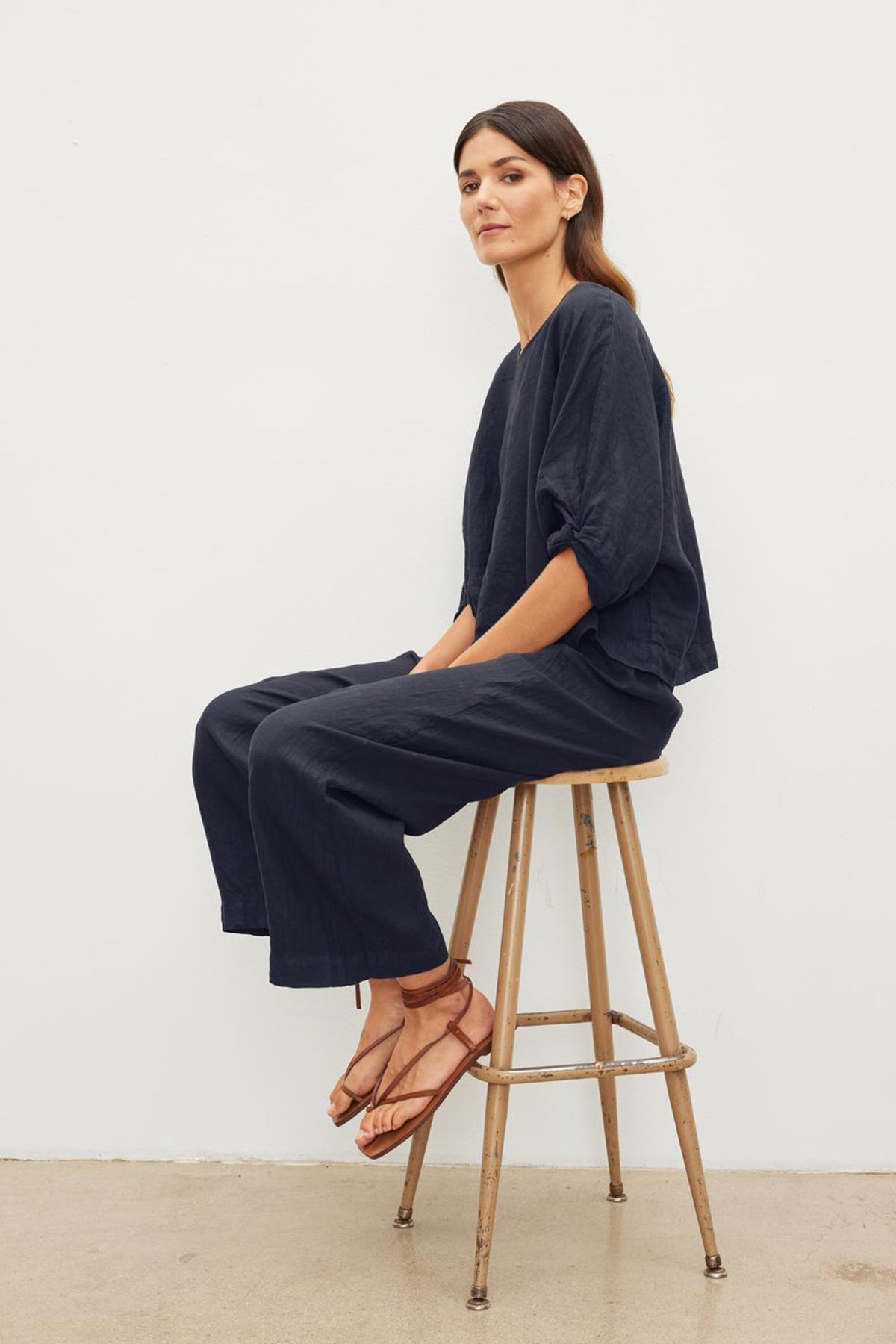 Woman with long dark hair sits on a wooden stool wearing a loose-fitting navy outfit with Velvet by Graham & Spencer's LOLA LINEN PANT and brown sandals, against a plain white background.-37240066441409