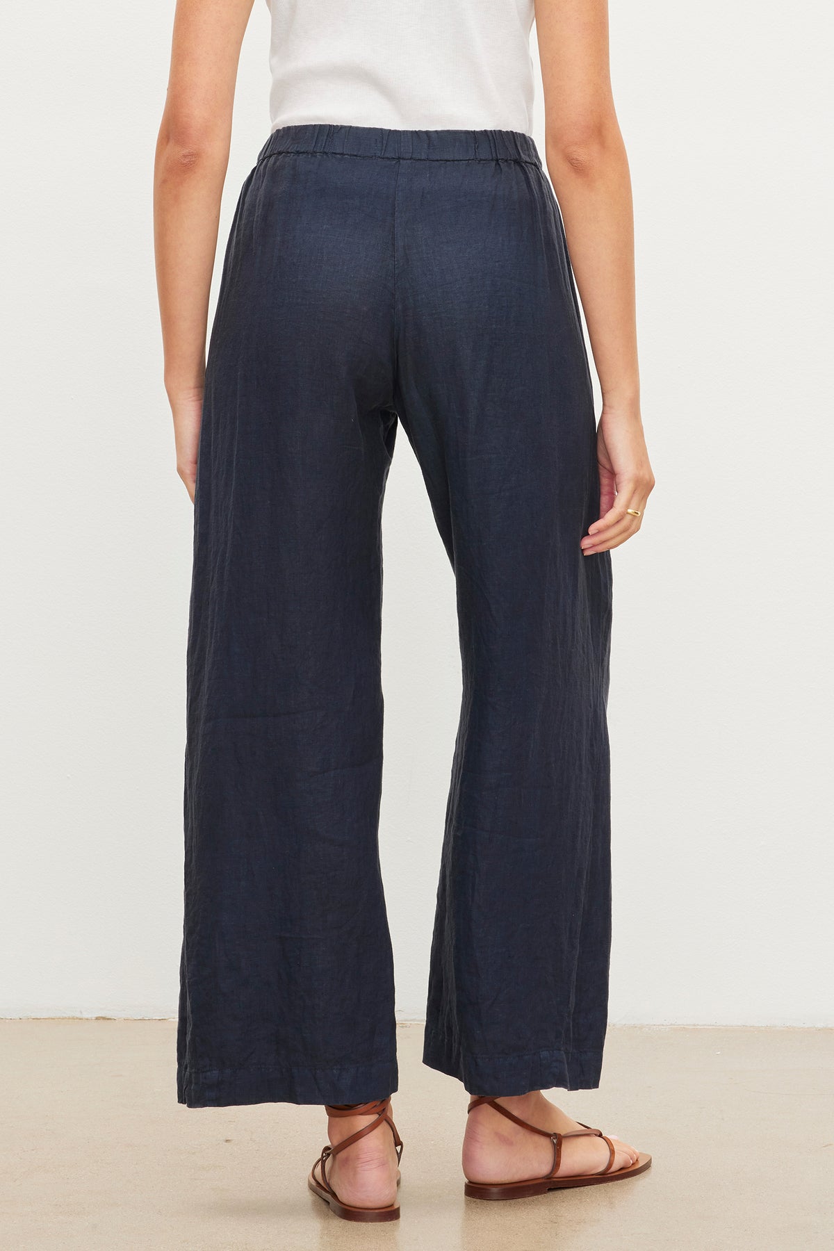   Woman standing, viewed from behind, wearing Loose Navy Blue LOLA LINEN PANT with an elastic waist and brown sandals by Velvet by Graham & Spencer. 