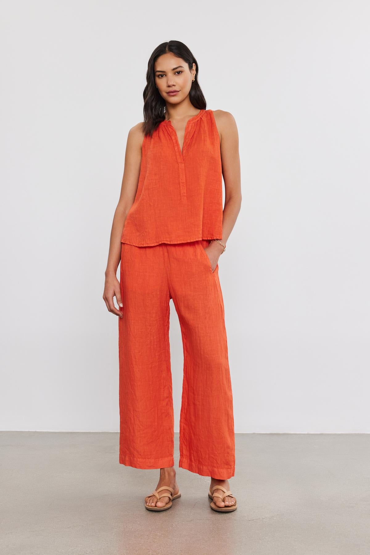   Person is standing against a plain background, wearing an orange sleeveless top and matching LOLA LINEN PANT by Velvet by Graham & Spencer. They are also wearing sandals. 