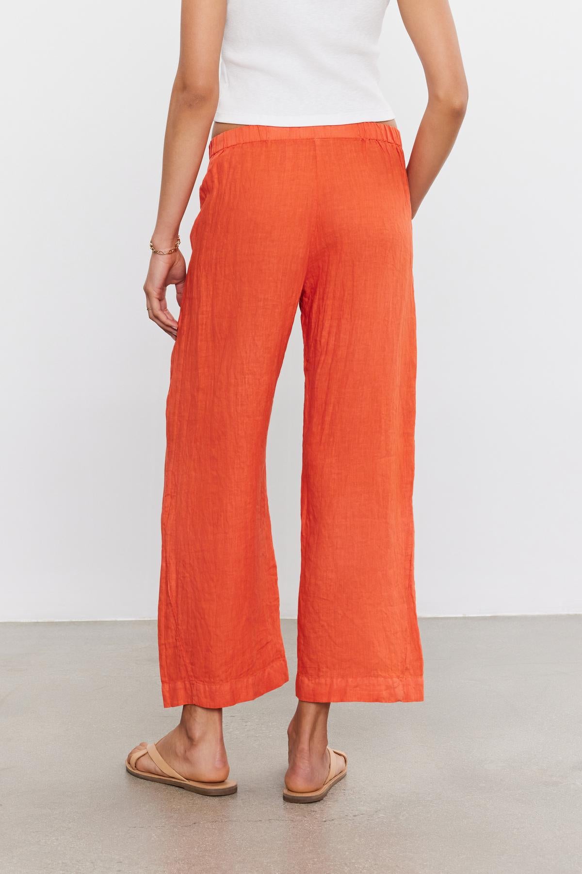   A person is standing, facing away, wearing bright orange LOLA LINEN PANT by Velvet by Graham & Spencer paired with a white top and beige flip-flops. 