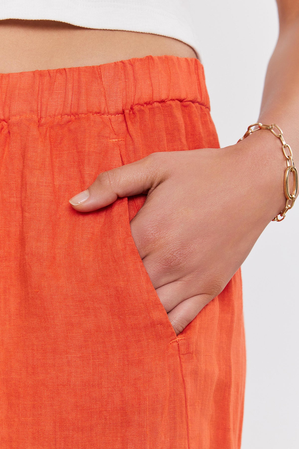 A person wearing relaxed leg, orange LOLA LINEN PANT by Velvet by Graham & Spencer with an elastic waistband has their hand in their pocket. They are also sporting a gold chain bracelet on their wrist.-36910043988161