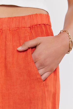 A person wearing relaxed leg, orange LOLA LINEN PANT by Velvet by Graham & Spencer with an elastic waistband has their hand in their pocket. They are also sporting a gold chain bracelet on their wrist.