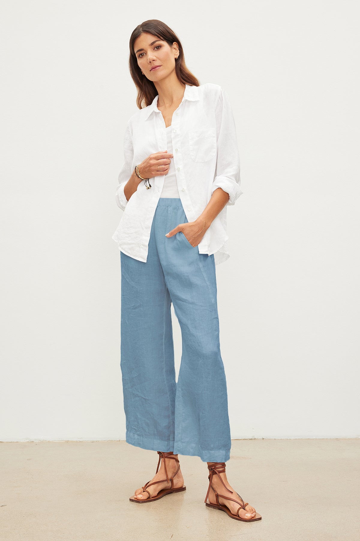 Woman standing with one hand in pocket, wearing a white shirt and blue LOLA LINEN PANT by Velvet by Graham & Spencer. She is also wearing brown strappy sandals and posing against a plain white background.-36161404895425