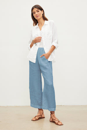 Woman standing with one hand in pocket, wearing a white shirt and blue LOLA LINEN PANT by Velvet by Graham & Spencer. She is also wearing brown strappy sandals and posing against a plain white background.