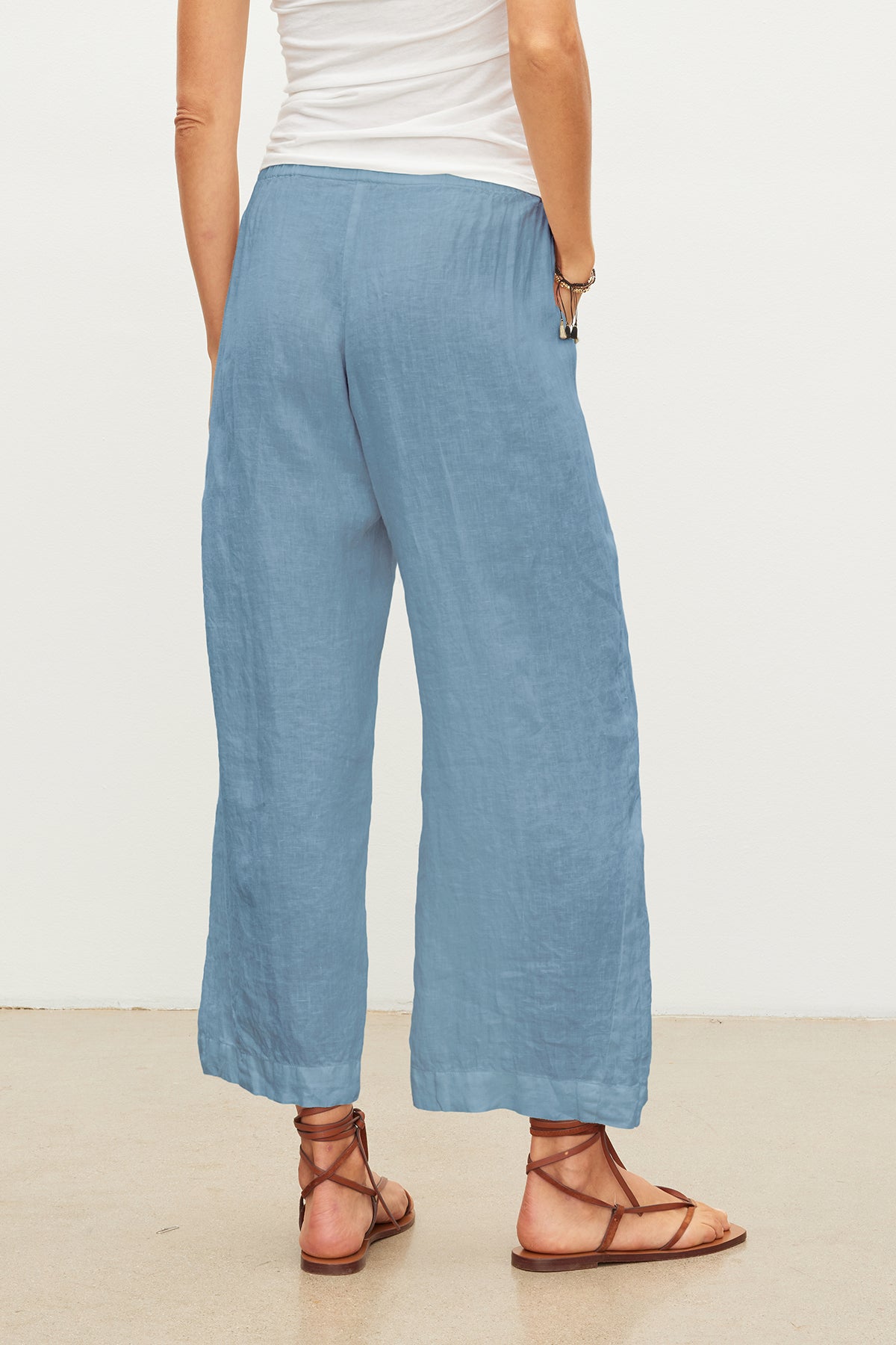   A person standing, wearing light blue wide-leg lightweight LOLA LINEN PANT by Velvet by Graham & Spencer, a white top, and brown sandals. The photo is taken from behind, showing the full length of the relaxed leg crops and sandals. 