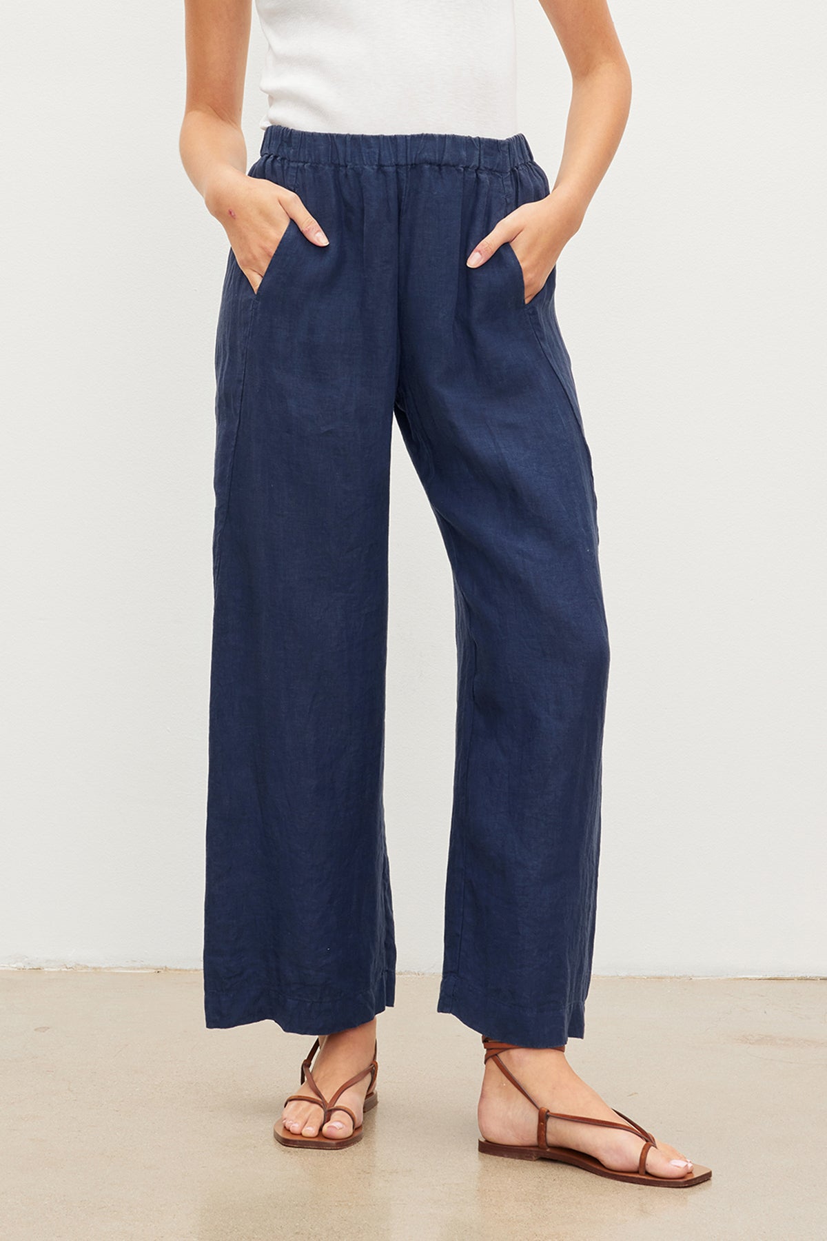 A person wearing a white top and navy blue LOLA LINEN PANT by Velvet by Graham & Spencer with hands in pockets. The individual also sports brown sandals and stands on a light-colored floor against a white wall.-36581057790145