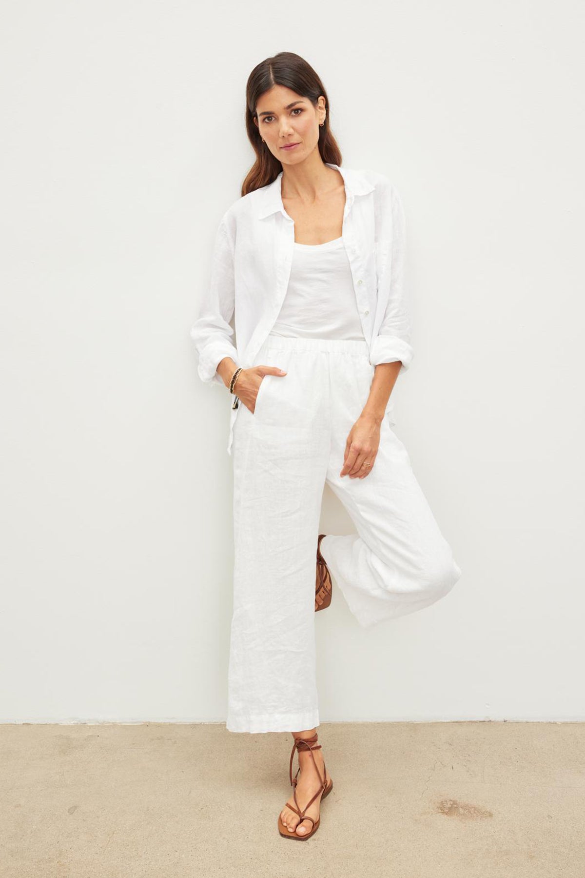   Woman in a white button-up shirt, white tank top, and Velvet by Graham & Spencer LOLA LINEN PANT stands against a plain wall. She is wearing brown sandals and has one hand in her pocket. 