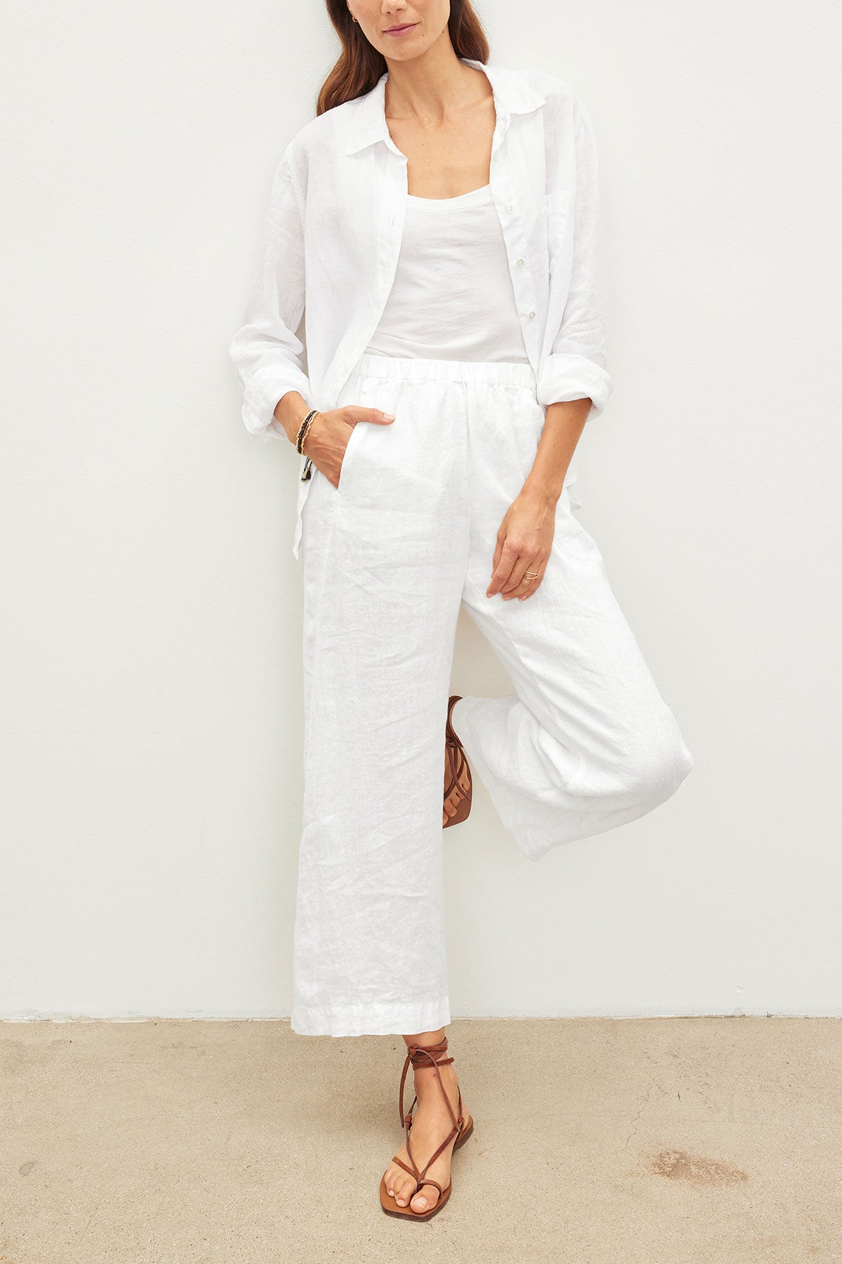 A woman stands against a white wall wearing a white shirt, white tank top, brown strappy sandals, and the LOLA LINEN PANT by Velvet by Graham & Spencer—lightweight pull-on trousers with an elastic waist—one leg bent, hand in pocket.-39418817413313