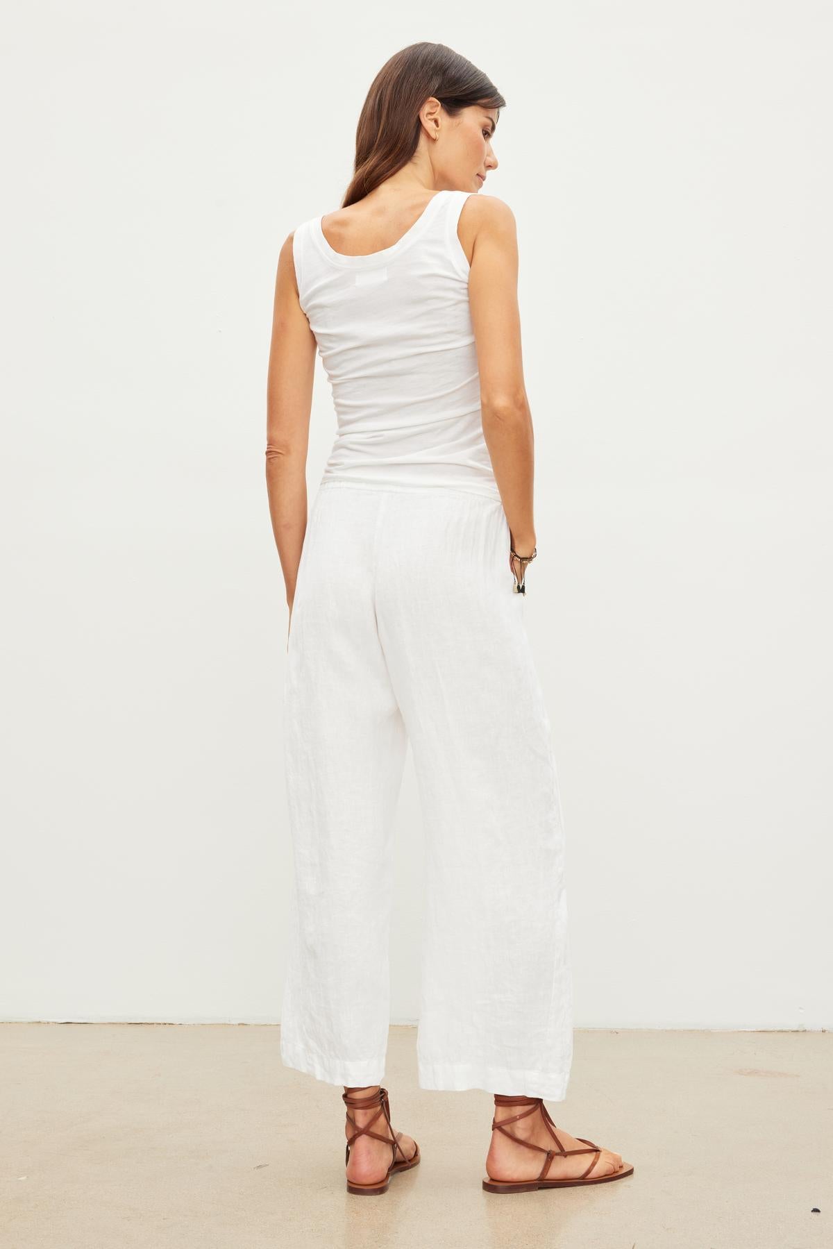 A woman stands facing away from the camera, wearing a white sleeveless top, LOLA LINEN PANT by Velvet by Graham & Spencer, and brown sandals. She is positioned against a plain white background.-36161692664001