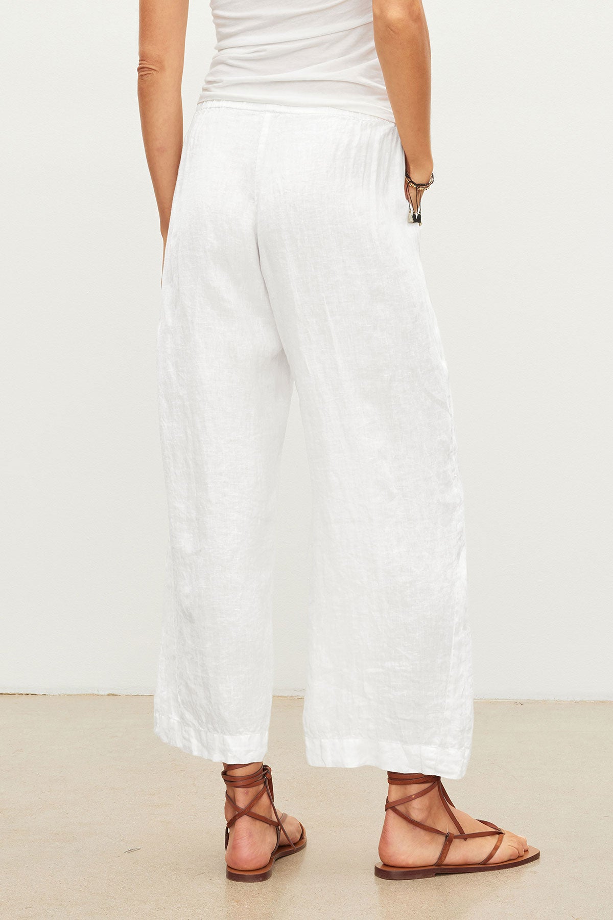 A person stands on a beige floor, seen from the back, wearing a white sleeveless top and Velvet by Graham & Spencer's LOLA LINEN PANT—lightweight linen pull-on trousers with relaxed legs—paired with brown strappy sandals.-39418729005249