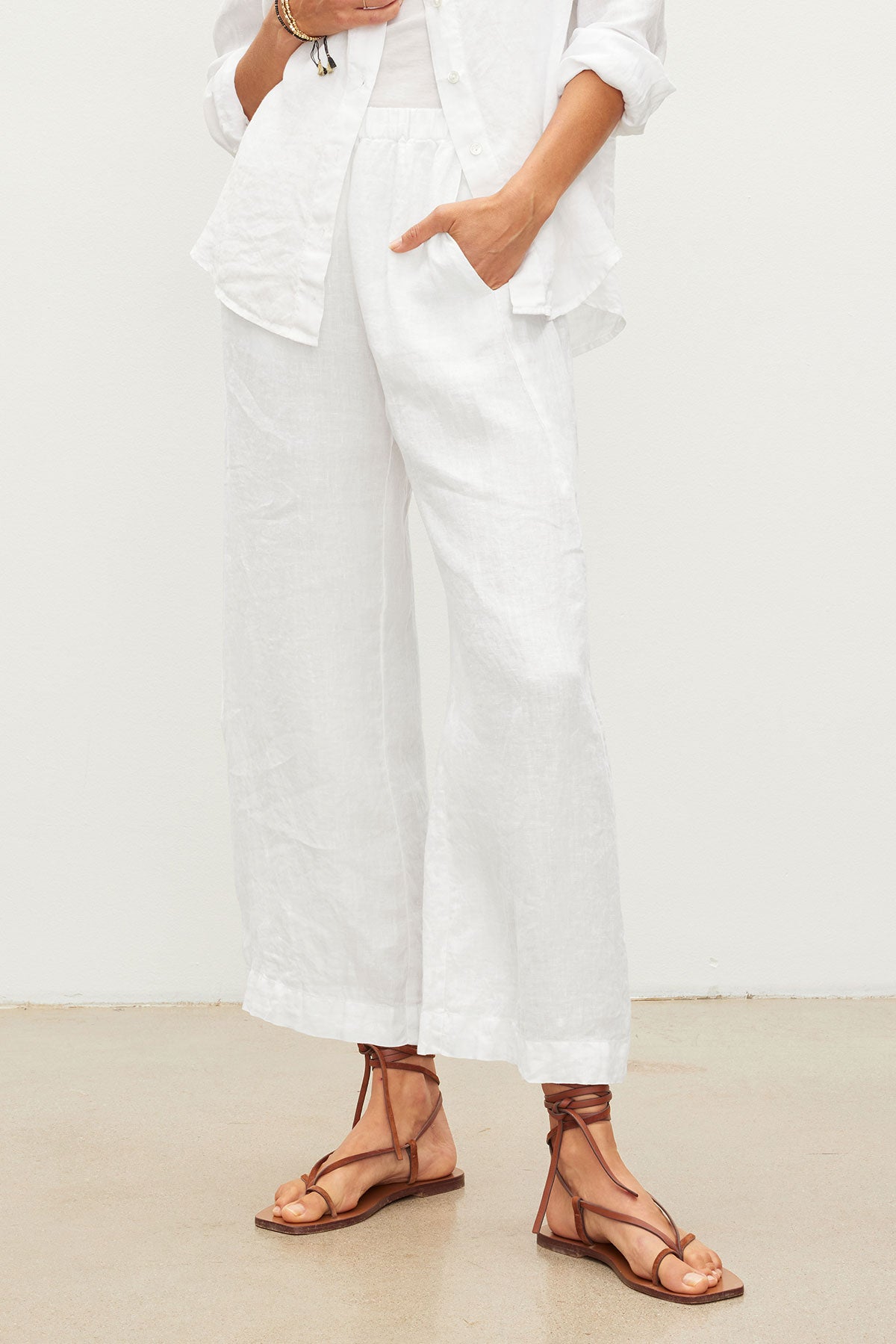  Model wearing the LOLA LINEN PANT by Velvet by Graham & Spencer, paired with a white button-up shirt and brown strappy sandals, standing against a plain light background. 