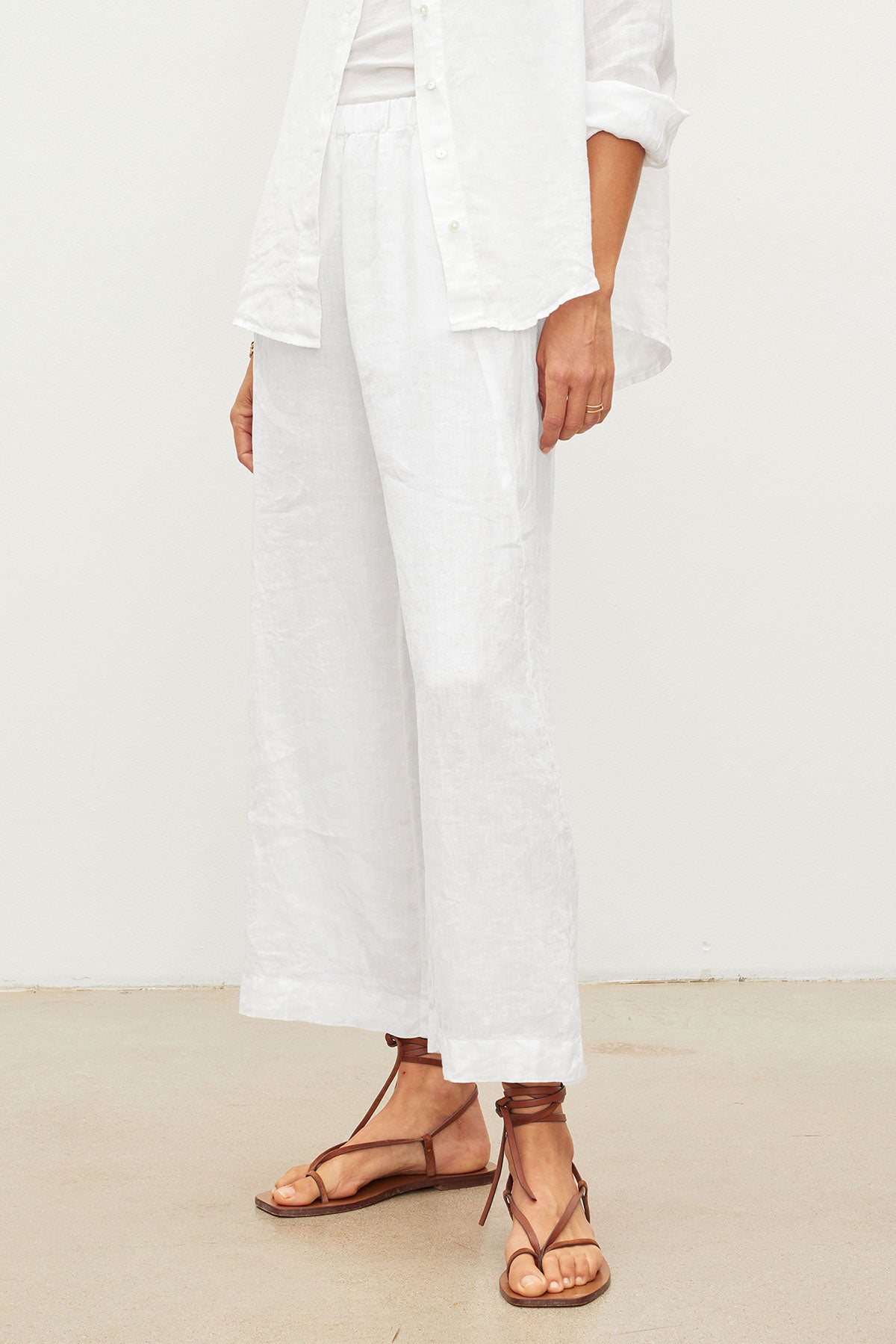 A person stands against a light background wearing a white button-up shirt, Velvet by Graham & Spencer's LOLA LINEN PANT with a relaxed leg, and brown lace-up sandals.-39418729038017