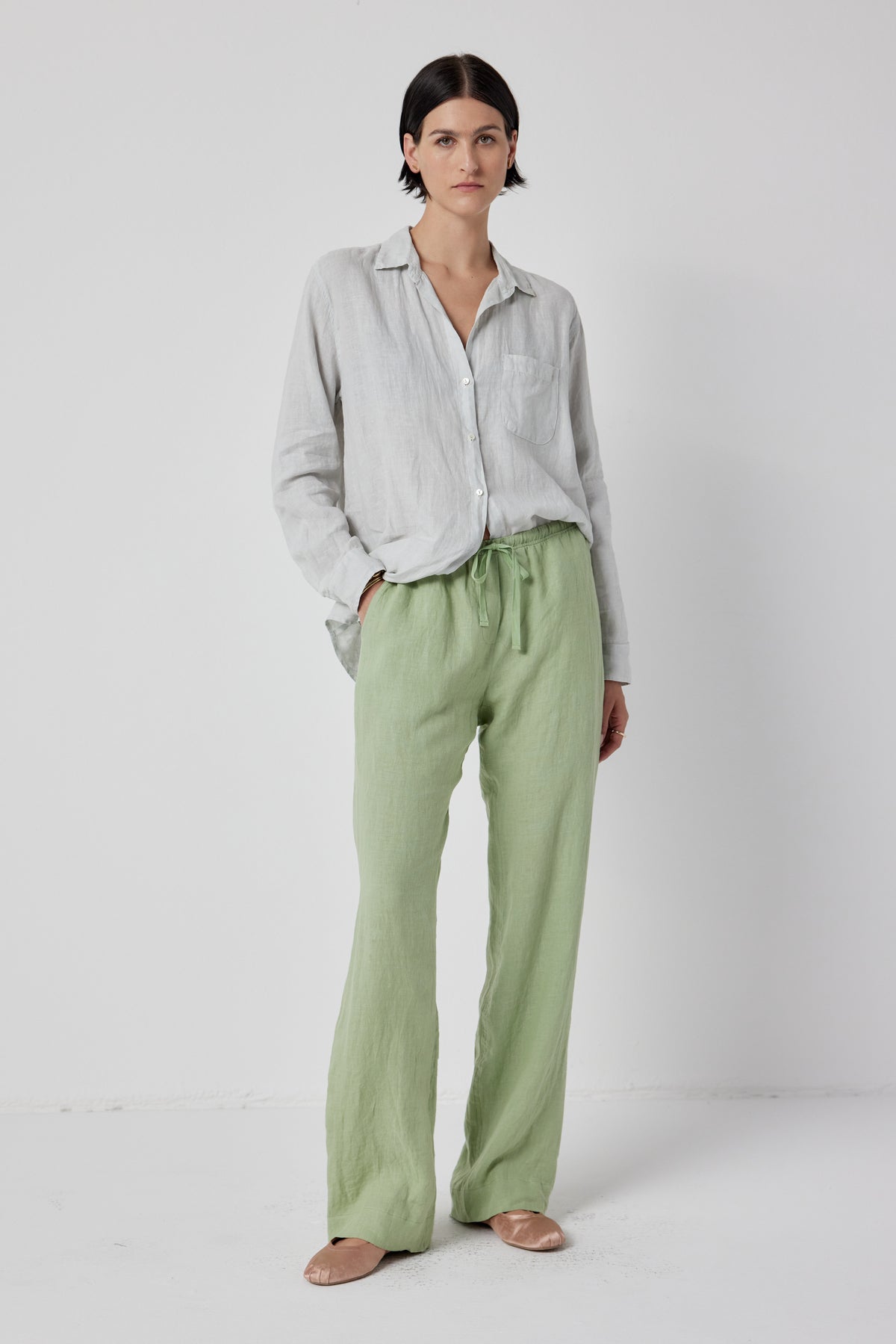   A woman stands against a white background wearing a loose-fitting grey shirt, soft linen Velvet by Jenny Graham PICO PANT, and beige flat shoes. 