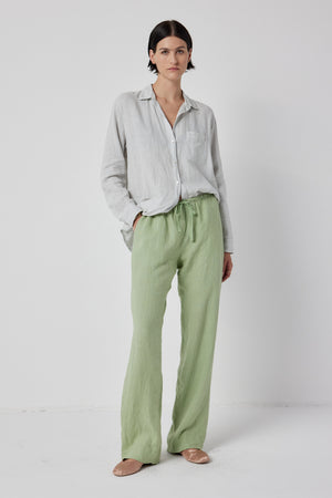 A woman stands in a studio wearing a light gray shirt, Velvet by Jenny Graham PICO LINEN PANT, and tan loafers, with hands partially in pockets, looking directly at the camera.
