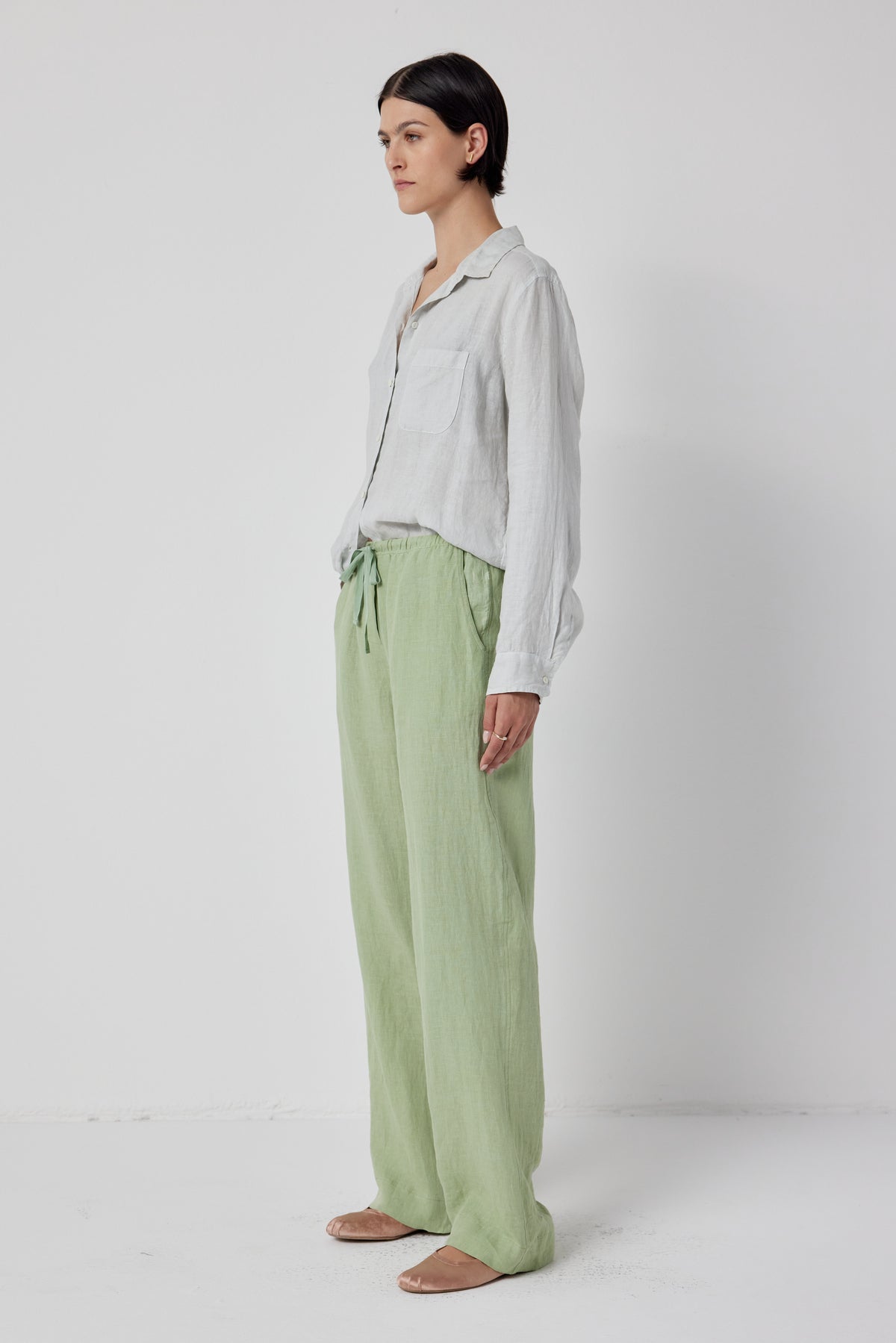   Woman standing in a studio, wearing a casual white shirt and light green Velvet by Jenny Graham Pico Linen Pant with an elastic waist, looking to the side, with a neutral expression. 