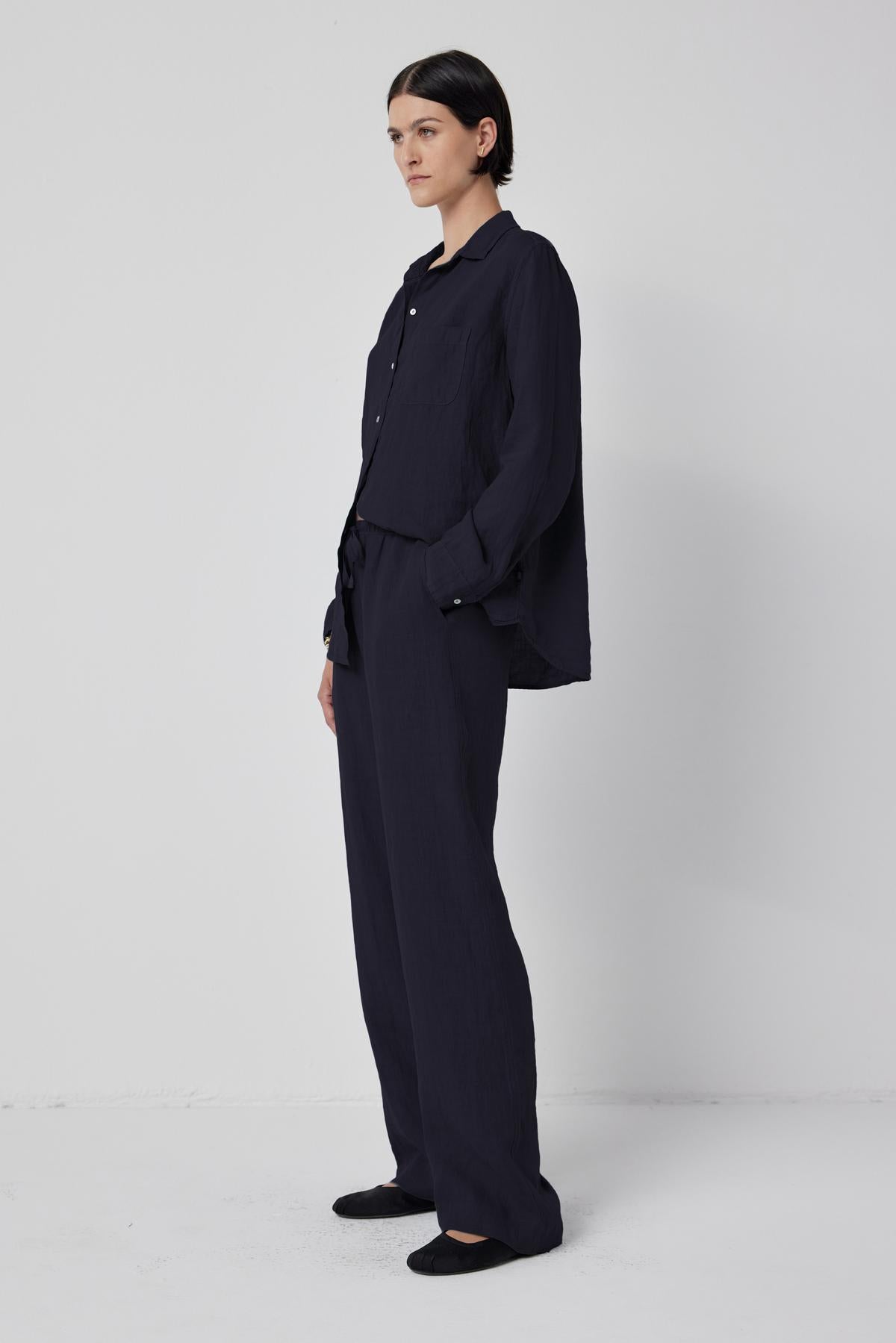   A person is standing against a plain white background, wearing a black long-sleeve button-up shirt and Velvet by Jenny Graham's PICO LINEN PANT, featuring a relaxed fit and an elastic waist. 