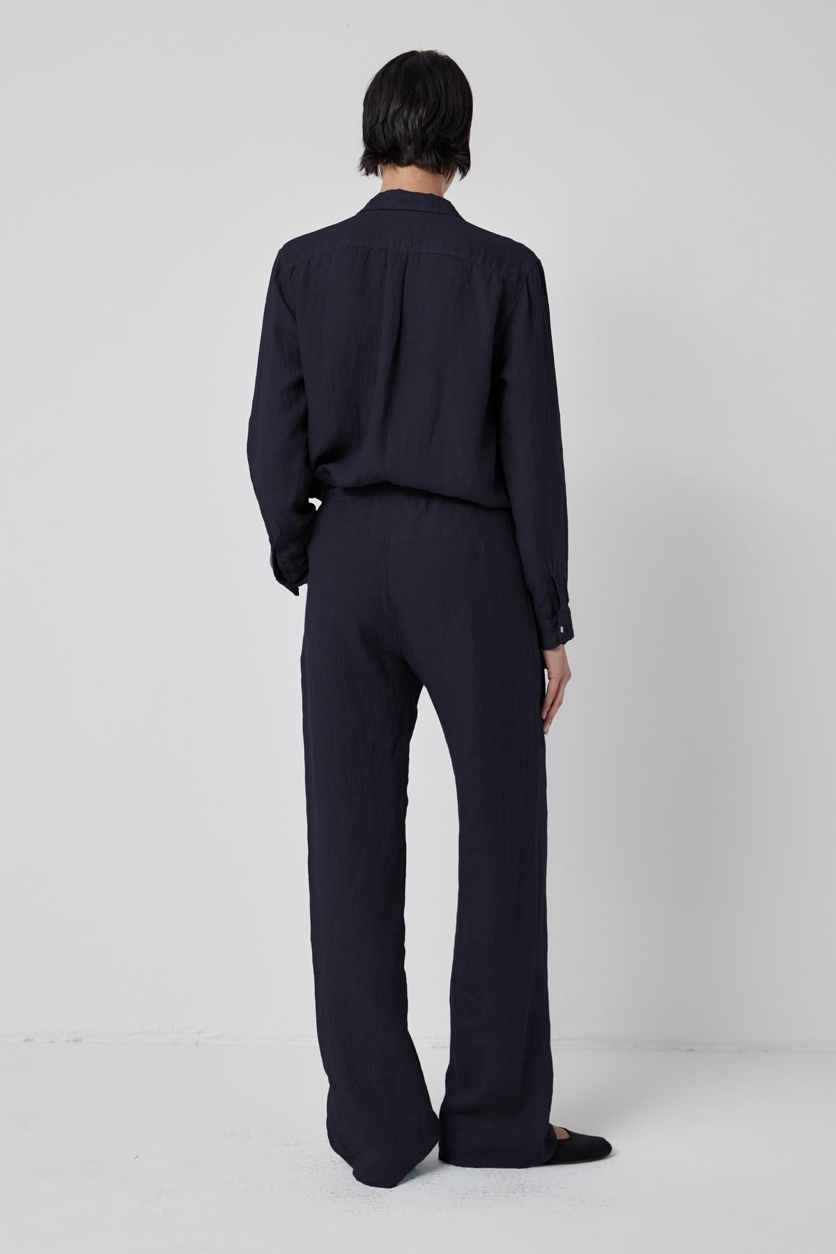   A person with short dark hair is standing with their back facing the camera. They are wearing a dark, long-sleeved shirt paired with the PICO LINEN PANT from Velvet by Jenny Graham, which has a matching relaxed fit. The background is plain and light-colored. 