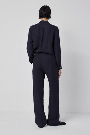 A person with short dark hair is standing with their back facing the camera. They are wearing a dark, long-sleeved shirt paired with the PICO LINEN PANT from Velvet by Jenny Graham, which has a matching relaxed fit. The background is plain and light-colored.