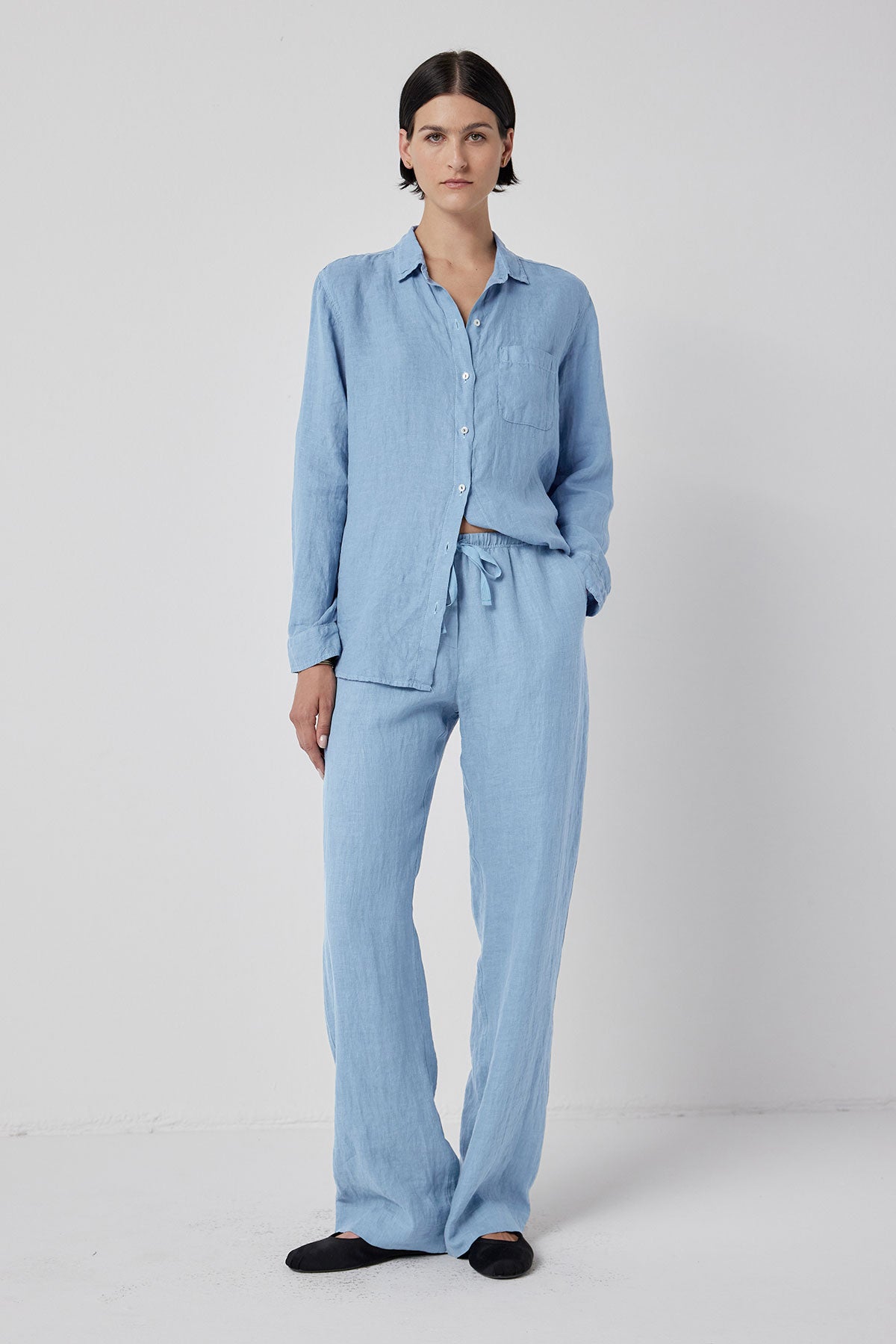   A woman standing in a studio wearing a light blue button-up shirt and matching PICO LINEN PANTS by Velvet by Jenny Graham, with her hands slightly tucked in relaxed fit linen pants. 