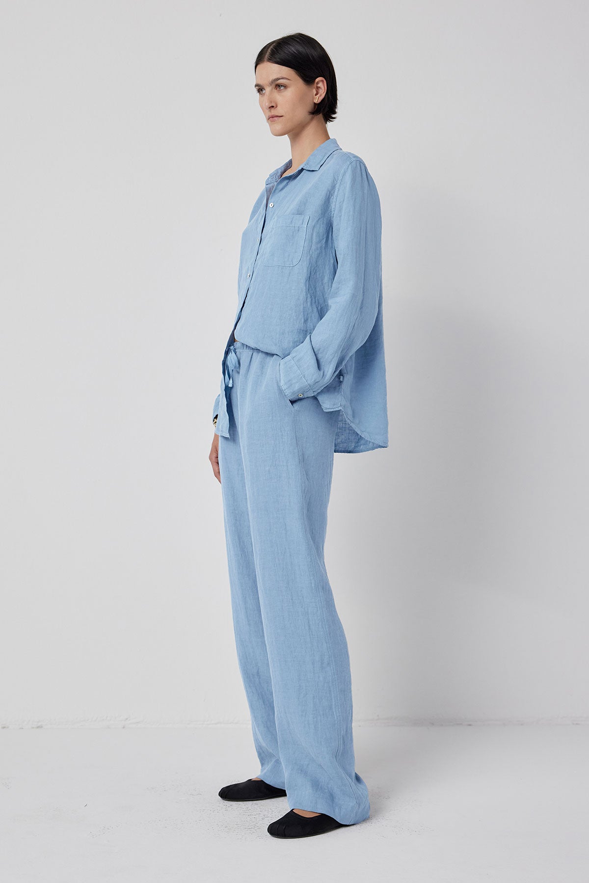   A woman standing and posing in a light blue linen shirt and matching PICO PANT trousers with an elastic waist drawstring against a white background by Velvet by Jenny Graham. 