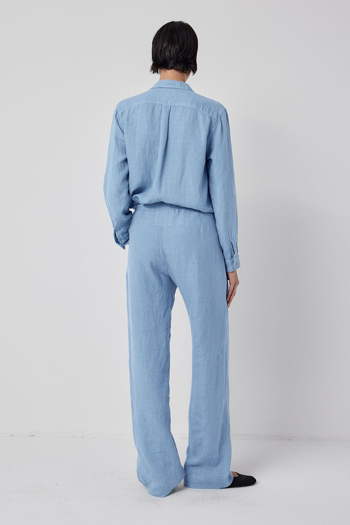 A person standing with their back to the camera, wearing a light blue denim jumpsuit with relaxed fit and black flats, against a white background.-36212497940673