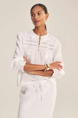 A woman stands with arms crossed against a plain light background, wearing the BYRDIE TOP by Velvet by Graham & Spencer—a romantic long-sleeve white embroidered cotton lace blouse—paired with a white skirt.