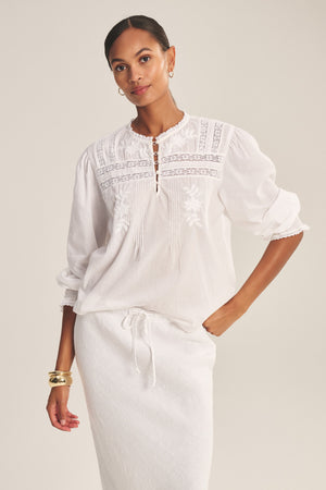 A woman wears the BYRDIE TOP by Velvet by Graham & Spencer—a white, long-sleeve embroidered blouse with a front keyhole—paired with a matching skirt, standing against a neutral background.