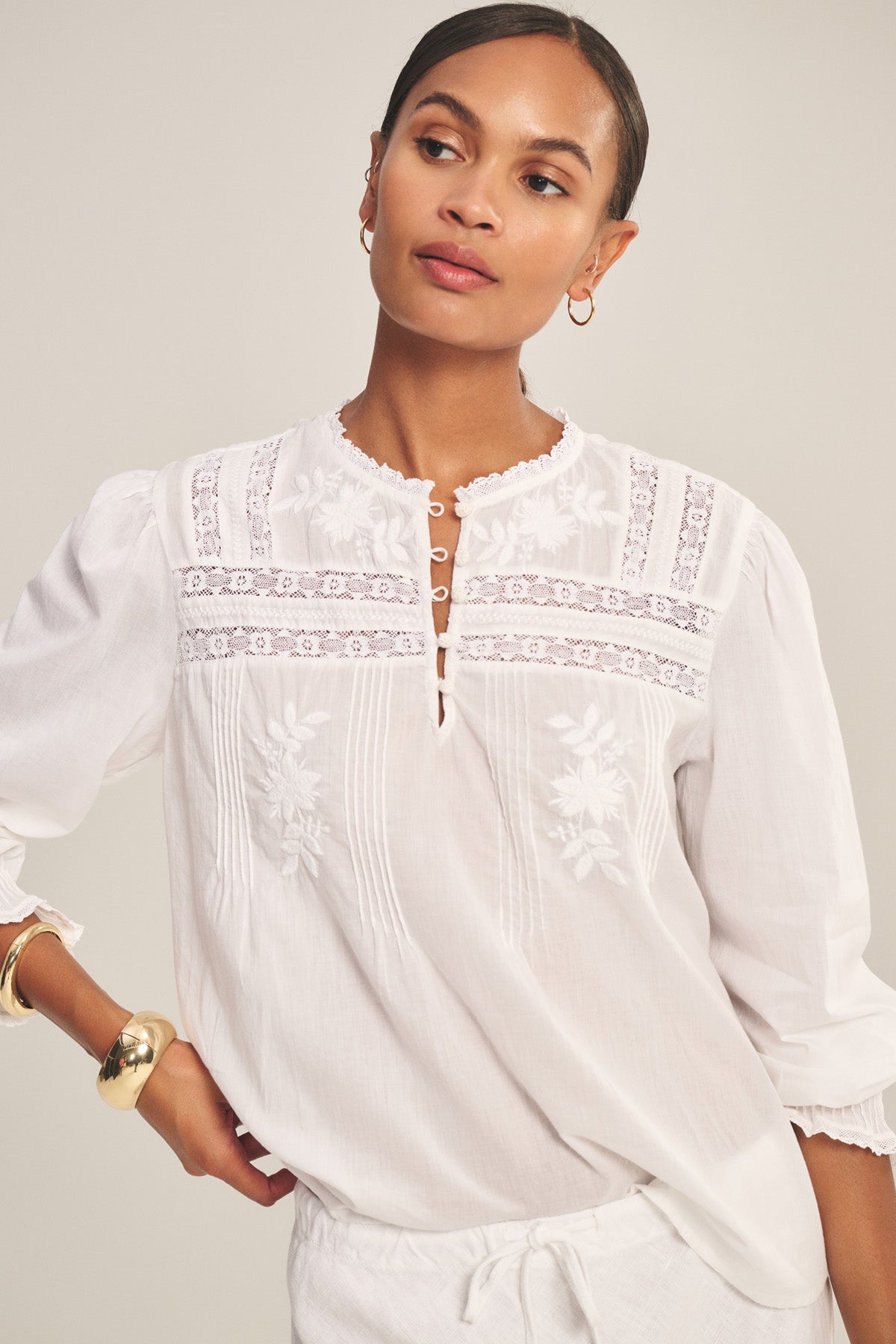 A woman wears the BYRDIE TOP by Velvet by Graham & Spencer—a white embroidered ruffle lace blouse—styled with gold jewelry, standing against a plain background.