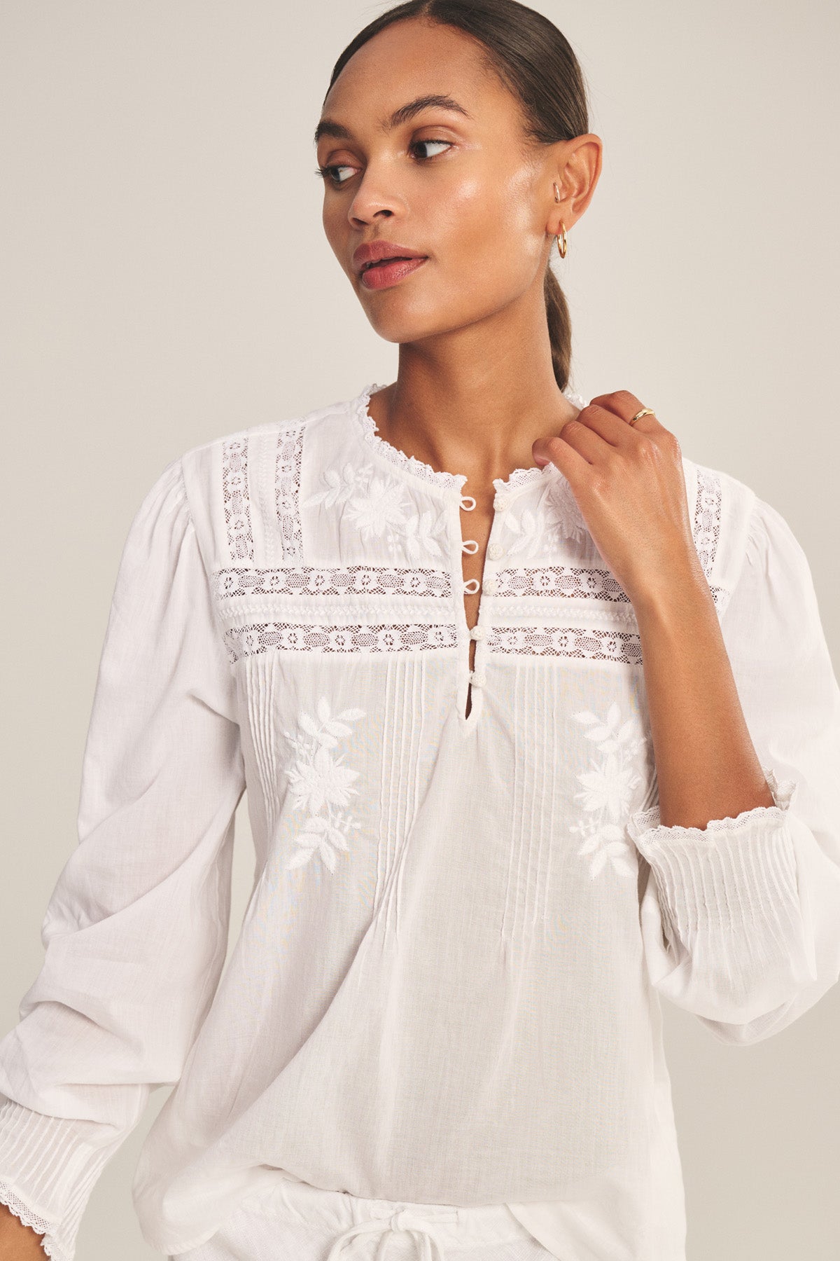 A woman wears the BYRDIE TOP by Velvet by Graham & Spencer—a white long-sleeve blouse with embroidered floral details and lace trim—standing against a light background.