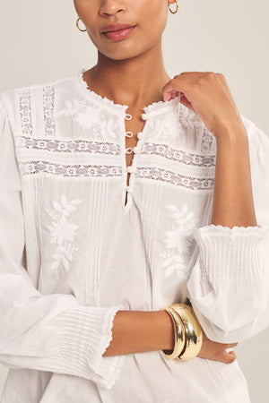 A woman wearing the BYRDIE TOP by Velvet by Graham & Spencer, featuring white embroidered details and gold bangles, stands against a plain background.