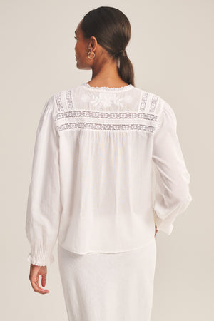 A woman with her hair in a low ponytail wears the BYRDIE TOP by Velvet by Graham & Spencer, featuring lace detailing on the back and long sleeves, paired with a white skirt.