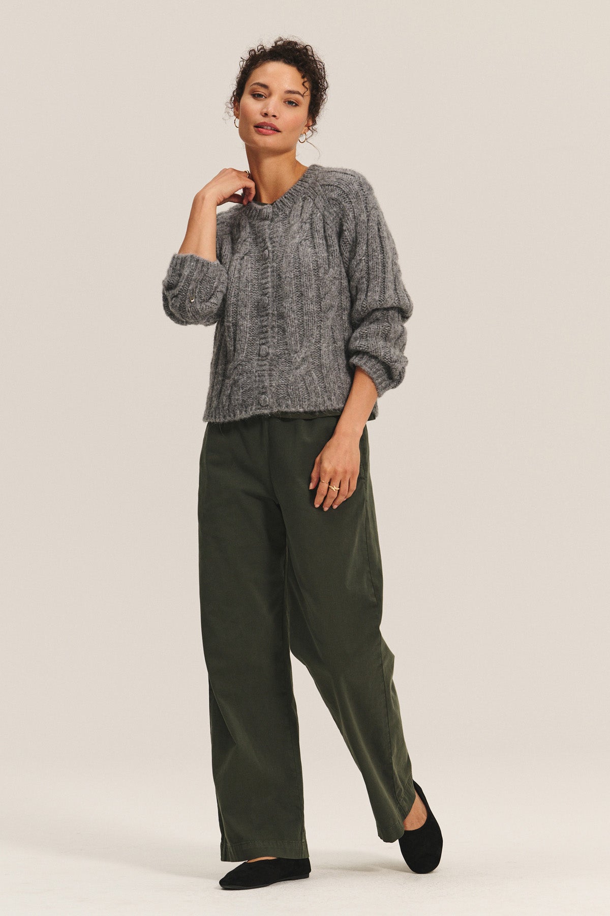 A woman stands against a plain background, layering the cozy Velvet by Graham & Spencer ALLISON CARDIGAN in grey over green wide-leg pants and black shoes.