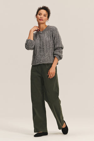 A woman stands against a plain background, layering the cozy Velvet by Graham & Spencer ALLISON CARDIGAN in grey over green wide-leg pants and black shoes.