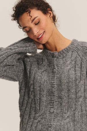 A woman in the ALLISON CARDIGAN by Velvet by Graham & Spencer smiles with her eyes closed, touching her hair and embracing the cozy charm of this gray cable-knit layering piece.