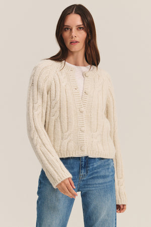 A woman in the ALLISON CARDIGAN by Velvet by Graham & Spencer, layered over a white shirt and blue jeans, stands against a plain background.