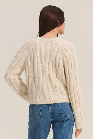 A woman with long brown hair is seen from the back wearing the Velvet by Graham & Spencer ALLISON CARDIGAN, an alpaca blend piece, paired with blue jeans against a plain background.