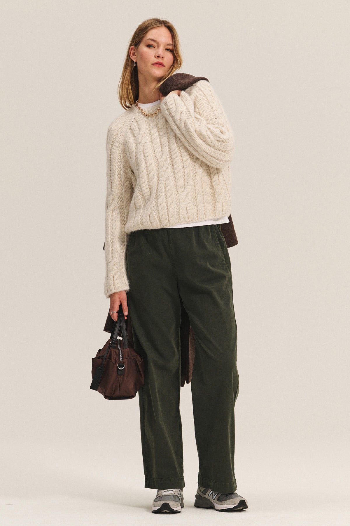 A woman wears the MARINA SWEATER by Velvet by Graham & Spencer—a cream cable knit with metallic threading—paired with dark green pants, gray sneakers, a brown bag, and a brown coat over her shoulder, standing against a plain background.-40248179458241
