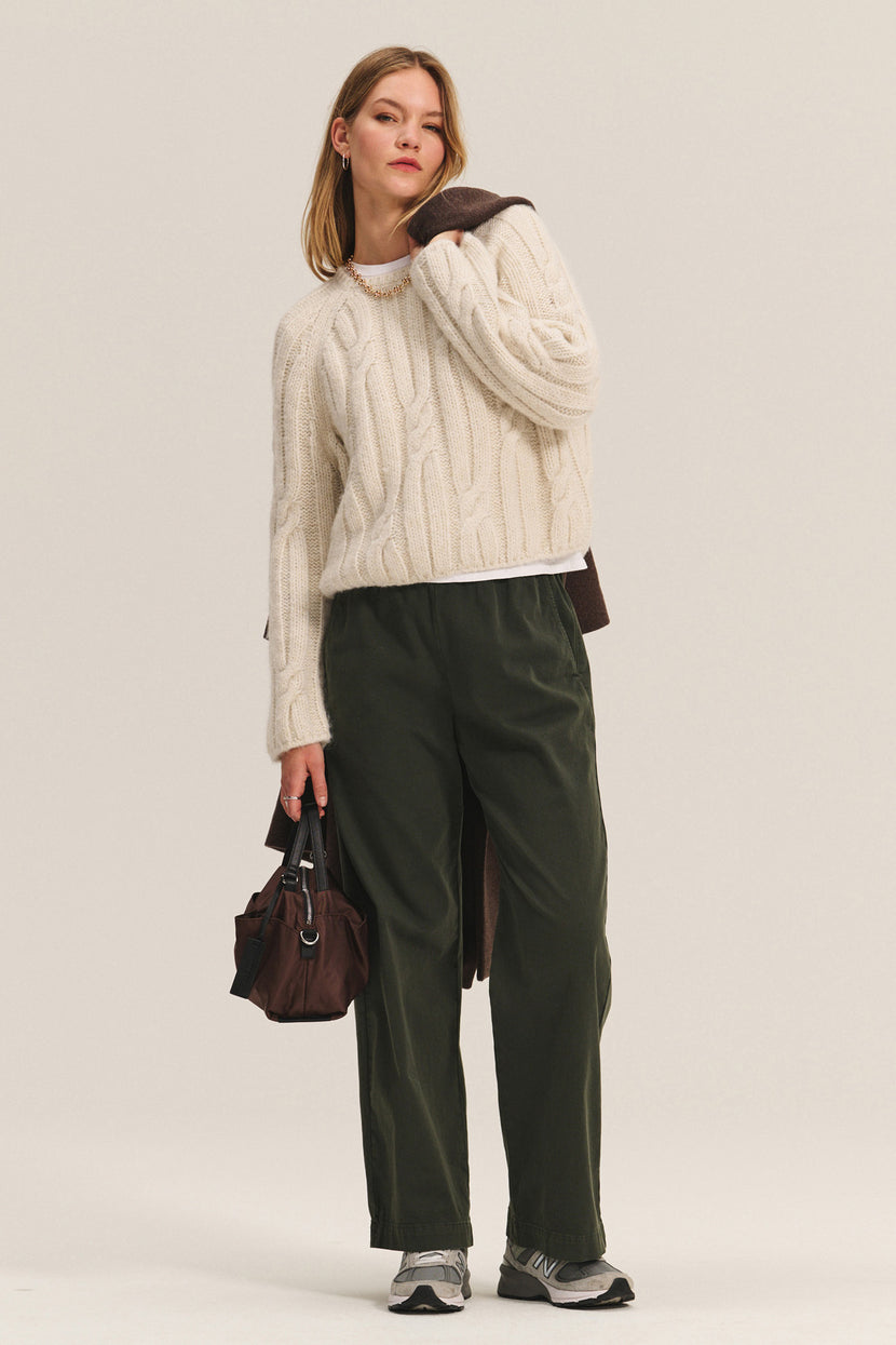 A woman wears the MARINA SWEATER by Velvet by Graham & Spencer—a cream cable knit with metallic threading—paired with dark green pants, gray sneakers, a brown bag, and a brown coat over her shoulder, standing against a plain background.