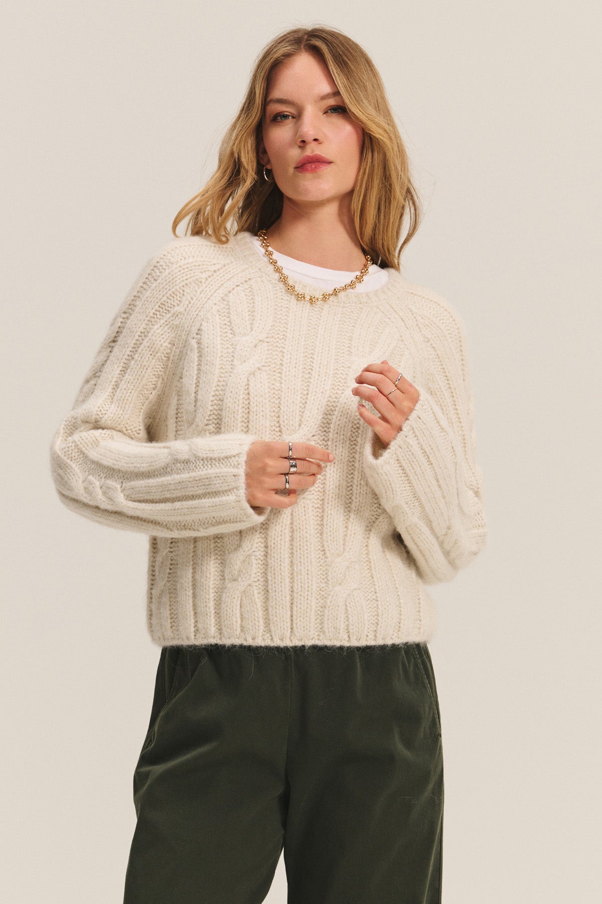 A woman wears the MARINA SWEATER by Velvet by Graham & Spencer—a cream cable knit with subtle metallic threading—paired with dark green pants and jewelry, standing against a plain light background.-40248179359937