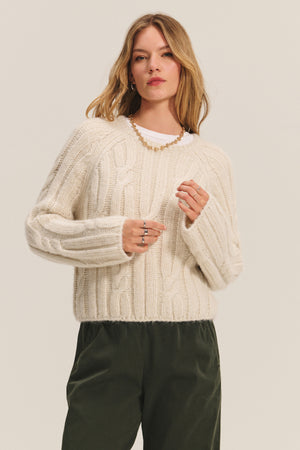 A woman wears the MARINA SWEATER by Velvet by Graham & Spencer—a cream cable knit with subtle metallic threading—paired with dark green pants and jewelry, standing against a plain light background.