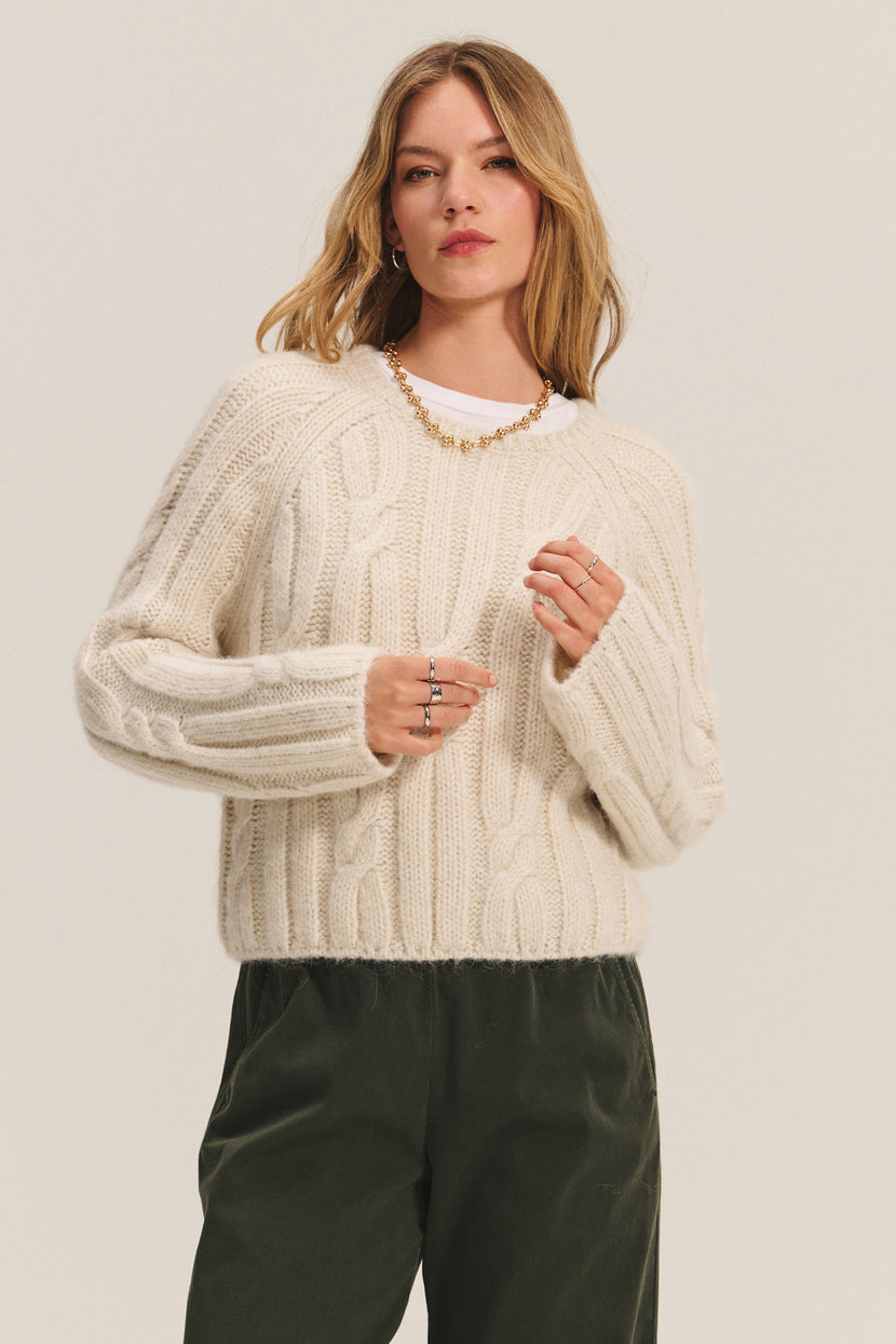 A woman wears the MARINA SWEATER by Velvet by Graham & Spencer—a cream cable knit with subtle metallic threading—paired with dark green pants and jewelry, standing against a plain light background.