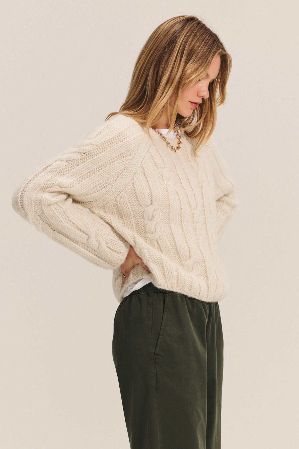 A woman wears the MARINA SWEATER by Velvet by Graham & Spencer—cream-colored, cable knit with metallic threading—and dark green pants, standing sideways with hands on hips against a light background.-40248179392705