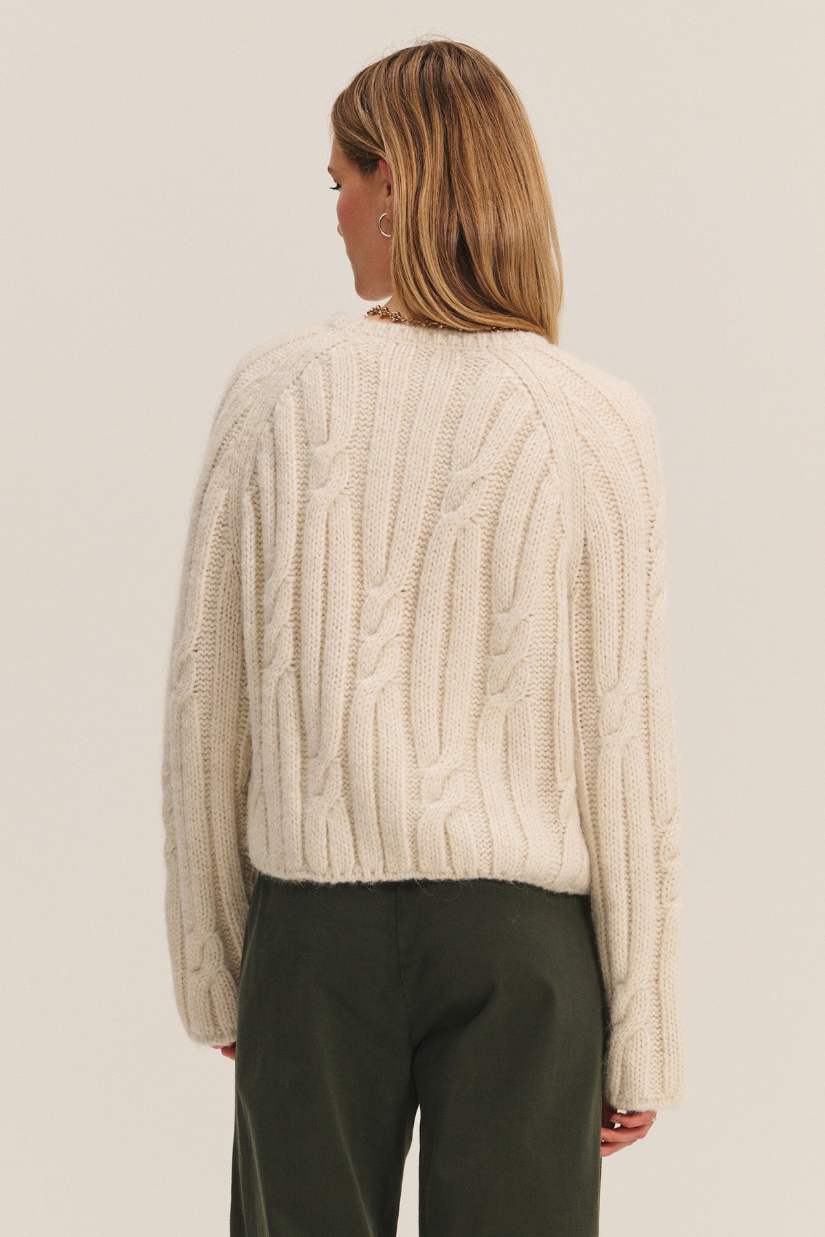 A woman with straight, light brown hair stands with her back to the camera, wearing Velvet by Graham & Spencer's MARINA SWEATER—a cream-colored cable knit with subtle metallic threading—paired with dark green pants against a plain background.