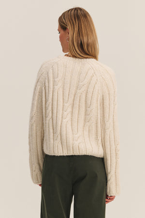 A woman with straight, light brown hair stands with her back to the camera, wearing Velvet by Graham & Spencer's MARINA SWEATER—a cream-colored cable knit with subtle metallic threading—paired with dark green pants against a plain background.